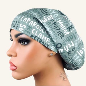 May include: A sage green scrub cap featuring a repeating pattern of white and dark green medical text including words like LAPAROSCOPE, CLAMP, RETRACT, MAYO, NIFE, SURGERY, FRAZIER, ADSON, and SCISSORS printed in various fonts and orientations across the fabric.