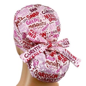 May include: A pink and white surgical scrub cap with a bow. The cap is covered in words like "Curette", "Needle Driver", and "Mosquito". The bow is tied at the back of the head. The cap is made of a soft fabric.