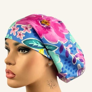May include: A scrub cap featuring a vibrant floral pattern with large pink, blue, and teal flowers on a light blue background. The fabric has a smooth texture and is styled in a bouffant shape that covers the head, with a wide band across the forehead.