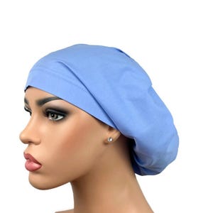 May include: A solid light blue scrub cap with a rounded, gathered crown and a wide, smooth band that fits snugly around the head. The fabric is a plain, matte cotton-like material in a soft sky blue shade, designed to cover the hair completely.