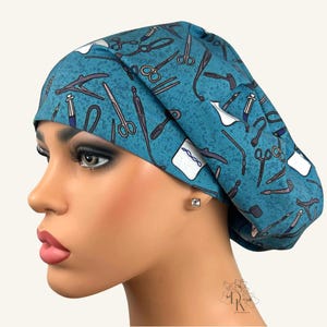May include: A teal scrub cap featuring a repeating pattern of various medical instruments, including scissors, forceps, and syringes, printed in dark blue and white. The fabric has a subtle textured appearance and a wide, folded front band.