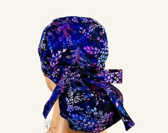 May include: A scrub cap featuring a dark blue background with a vibrant floral pattern in shades of purple, pink, and light blue. The cap has a rounded crown and a tie-back closure with two fabric tails that are knotted at the back of the head.