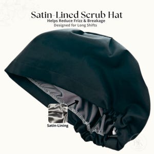 Op de afbeelding: Een zwarte operatiemuts met een grijze satijnen voering, voorzien van de tekst Satin-Lined Scrub Hat, Helps Reduce Frizz & Breakage, Designed for Long Shifts en Satin-Lining. De muts heeft een geplooide achterkant met een verstelbare sluiting.