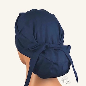 May include: A solid navy blue scrub cap featuring a gathered back design with fabric ties. The cap is constructed with a wide band that wraps around the head and a pleated pouch at the rear to accommodate hair, providing a secure and adjustable fit.
