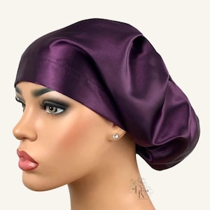 May include: A deep purple satin bonnet with a smooth, lustrous finish and a wide, structured band that fits securely around the head. The fabric features soft, gathered folds at the back, creating a voluminous, rounded shape for hair coverage.