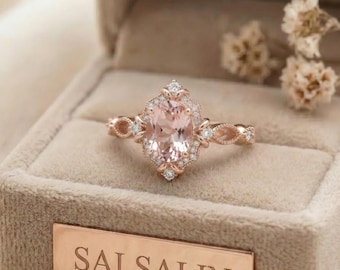 May include: A rose gold ring featuring an oval-cut peach-colored center stone surrounded by a halo of small white stones. The band has an intricate openwork design. The ring is displayed in a beige velvet box with the text SALSALIN on a plate.