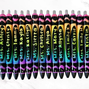 May include: A collection of personalized pens with names like "Mrs. Kristan" and "Ms. Lindsey" in a variety of colors. The pens have a black barrel with a glittery rainbow design and a silver tip. The names are written in a handwritten style.