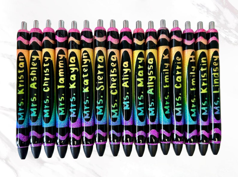 May include: A collection of personalized pens with names like "Mrs. Kristan" and "Ms. Lindsey" in a variety of colors. The pens have a black barrel with a glittery rainbow design and a silver tip. The names are written in a handwritten style.
