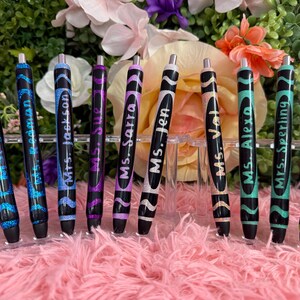 May include: A collection of black pens with glitter and wavy designs. Each pen is labeled with a name in white script. The pens are displayed on a clear acrylic stand against a floral background.