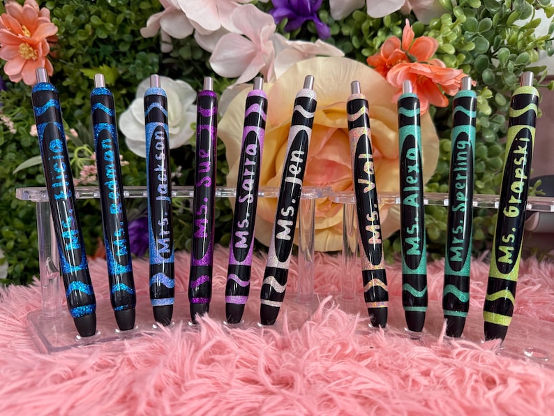 May include: A collection of black pens with glitter and wavy designs. Each pen is labeled with a name in white script. The pens are displayed on a clear acrylic stand against a floral background.