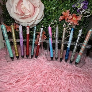 May include: A selection of glitter pens in a range of colours, such as teal, gold, purple, red, and blue, presented on a clear acrylic stand. The pens have a shiny, textured finish and are arranged in a row. The background includes flowers and a pink, fluffy surface.