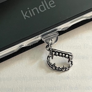 May include: A silver charm in the shape of a set of fangs, hanging from a black e-reader with the word "kindle" on the front.
