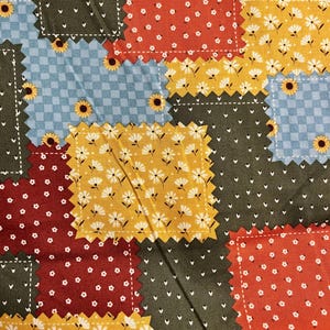 Pode incluir: Um edredom de retalhos com uma variedade de padrões de tecido. O edredom apresenta quadrados de tecido vermelho, amarelo, verde e azul. Alguns quadrados têm padrões de margaridas brancas, girassóis ou pequenos pontos brancos. As bordas são costuradas com fio branco.