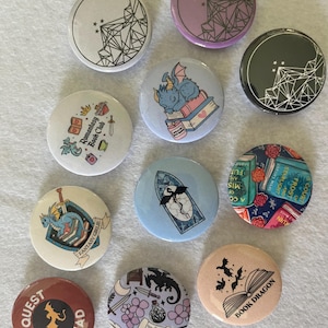 Puede incluir: Una colección de botones redondos con diseños de temática fantástica. Los botones presentan ilustraciones de dragones, libros, formas geométricas y textos como "Quest Squad" y "Book Dragon". La paleta de colores incluye azules, morados y marrones.