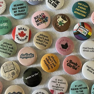 Może przedstawiać: Kolekcja kolorowych, okrągłych przypinek z różnymi frazami i grafikami związanymi z książkami. Przypinki zawierają teksty takie jak "Czytaj więcej książek", "Klub książki" i "Czytanie zrujnowało mój harmonogram snu". Niektóre przypinki mają ilustracje książek i filiżanek kawy.
