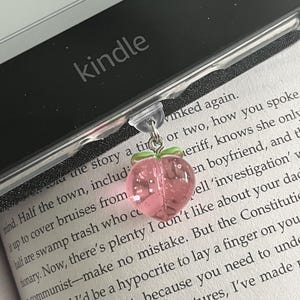 May include: A pink glass peach charm bookmark with a silver chain attached to a black Kindle e-reader. The Kindle is open to a page with text about a story and a town.