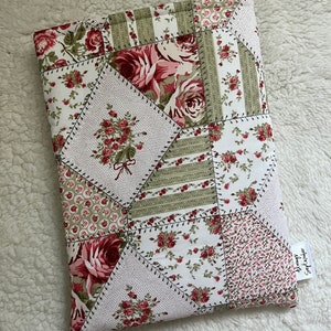 Puede incluir: Una funda de libro de tela de retazos con varios patrones florales en tonos rojos, rosas y verdes sobre un fondo blanco. La funda está hecha de piezas de tela cosidas y tiene una pequeña etiqueta con texto.