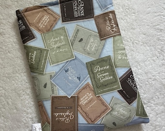 May include: A fabric book sleeve featuring a light blue background with a pattern of book covers. The book covers are in shades of brown, green, and blue, with titles like "Anne of Green Gables" and "Rilla of Ingleside". A small white tag reads "Always September".