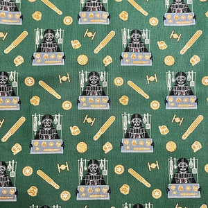 Puede incluir: Una tela verde con un patrón repetido de Darth Vader sosteniendo una bandeja de galletas. Las galletas son de diferentes formas y tamaños. También hay otros elementos con temática de Star Wars en el patrón, como sables de luz y cazas TIE.