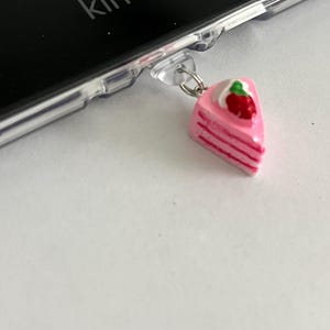 May include: A pink cake slice charm with a strawberry on top, attached to a clear phone case. The word "kindle" is visible on the black device.
