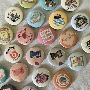 May include: A selection of colourful round badges with book-related designs and text. The badges feature illustrations of books, cats, dogs, and other related imagery. Some badges include phrases like "Happily Ever After" and "Read More Books."