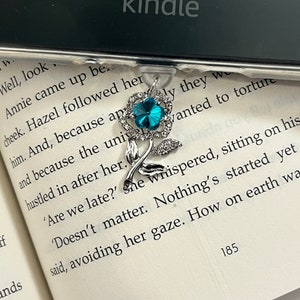 May include: A silver bookmark with a turquoise rhinestone flower design. The bookmark is attached to a clear plastic holder. The text "kindle" is visible on the top of the holder.