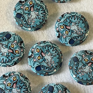 May include: A collection of round, blue cat-themed buttons. Each button features a cartoon cat with yellow eyes and its tongue sticking out. The cat is surrounded by a swirling pattern and small floral accents.