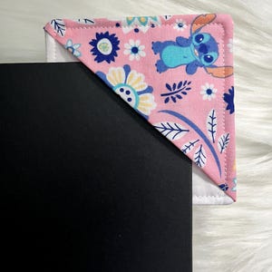 Puede incluir: Marcapáginas de tela rosa con un patrón floral y un personaje azul Stitch. El marcapáginas está doblado en forma de triángulo.