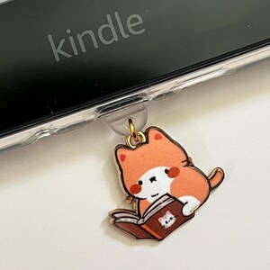 May include: A gold-coloured charm featuring a cartoon orange cat reading a book. The cat is sitting on a book with a white cat on the cover. The charm is attached to a clear plastic case with the word "kindle" on it.