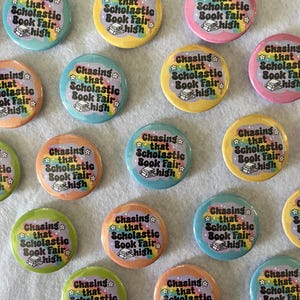 May include: A collection of colorful round buttons with the text "Chasing that Scholastic Book Fair high". The buttons feature a rainbow, stars, and a stack of books. Colors include pink, yellow, blue, orange, and green.