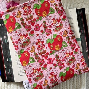 May include: Pink book sleeve with a repeating pattern of strawberries, houses, and a character named "Strawberry Shortcake". The sleeve has a white tag that says "Always September".