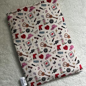 May include: A white laptop sleeve with a colourful pattern featuring the year 1989, lipstick, guitars, snakes, cowboy boots, and other 80s-inspired icons. The sleeve has a white tag with the text "Always September".