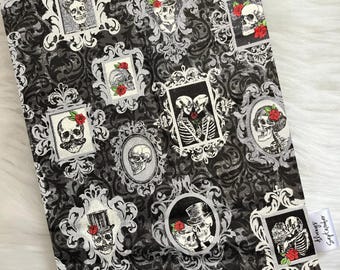 May include: A book cover with a black and white damask pattern, adorned with framed skull portraits and red roses. The cover includes a small white tag with text.