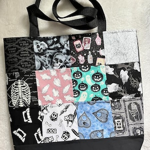 May include: A patchwork tote bag with a black base and straps. The bag's design features various Halloween-themed fabric squares, including skulls, ghosts, pumpkins, and Ouija board motifs. The bag is designed for carrying items.