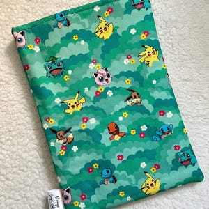 May include: Green fabric book sleeve featuring a repeating pattern of various Pokémon characters, including Pikachu, Bulbasaur, and Squirtle, set against a backdrop of stylized green clouds and flowers. The sleeve has a small white tag with the text "Always September".