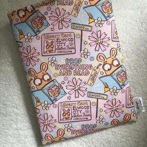May include: A padded book sleeve featuring a repeating pattern of the cartoon character Arthur reading a book, flowers, and library cards on a pink background. Text includes "Drop Everything and Read" and "Elwood City".