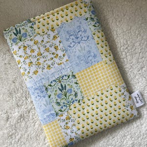 May include: A patchwork book cover in shades of yellow, blue, and white. The cover is made from various floral and patterned fabrics. It has a scalloped edge and a small tag with text. The cover is designed to protect books.