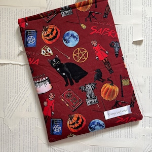 Puede incluir: Una cubierta de libro roja con un tema de Halloween que presenta un gato negro, calabazas, pentagramas y el texto "Chilling Adventures of Sabrina".