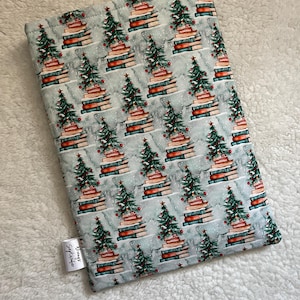 May include: A teal blue fabric book sleeve with a repeating pattern of Christmas trees made of stacked books. The trees are decorated with red ornaments and green garlands. The fabric has a subtle white snowflake pattern. The sleeve has a white tag that says "Sewing September".