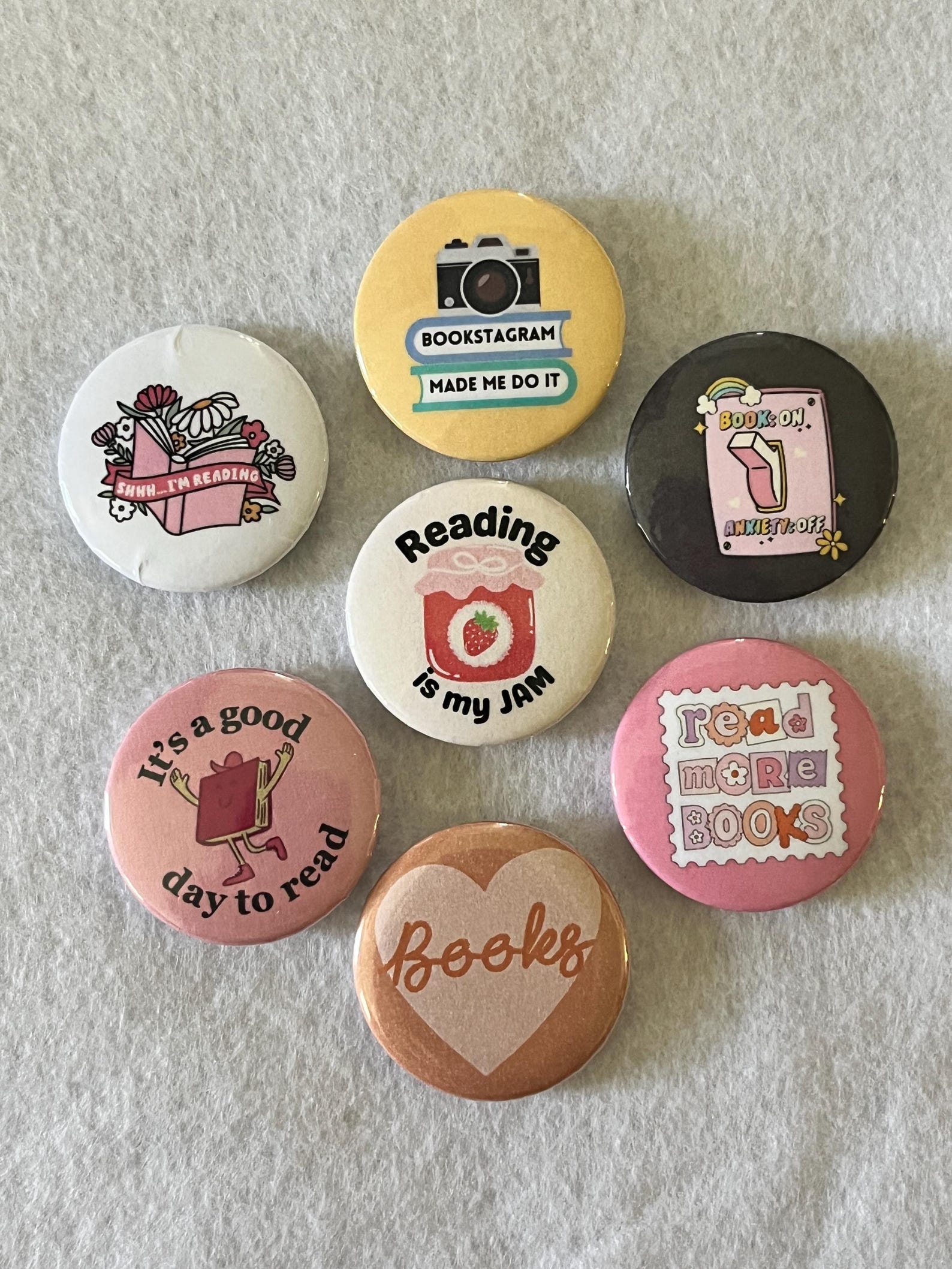 A collection of seven book-related pins with various sayings and illustrations on each one. Top left: A white pin with flowers and an open book, with a banner reading "Shhh... I’m reading.” Top center: A yellow pin featuring a camera and the words "Bookstagram made me do it.” Top right: A black pin showing a light switch with text "Book on, anxiety off.” Middle center: A white pin with a jam jar and the words "Reading is my JAM.” Bottom left: A pink pin with a happy book character and the phrase "It’s a good day to read.” Bottom center: A light brown pin with a heart background and the word "Books.” Bottom right: A pink pin with the words "Read more books" in a cut-out letter style.