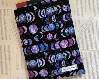 Peut inclure: Une housse de livre noire avec un motif répété de lunes et d'étoiles aquarelle violettes et bleues sur un fond noir. Le texte "Always September" est imprimé sur une étiquette blanche attachée à la couverture.