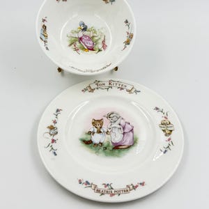 May include: A white ceramic bowl and plate set featuring illustrations from Beatrix Potter stories. The bowl has "Jemima Puddle Duck" text, and the plate has "Tom Kitten" and "Beatrix Potter" text. Both pieces have floral and character details.