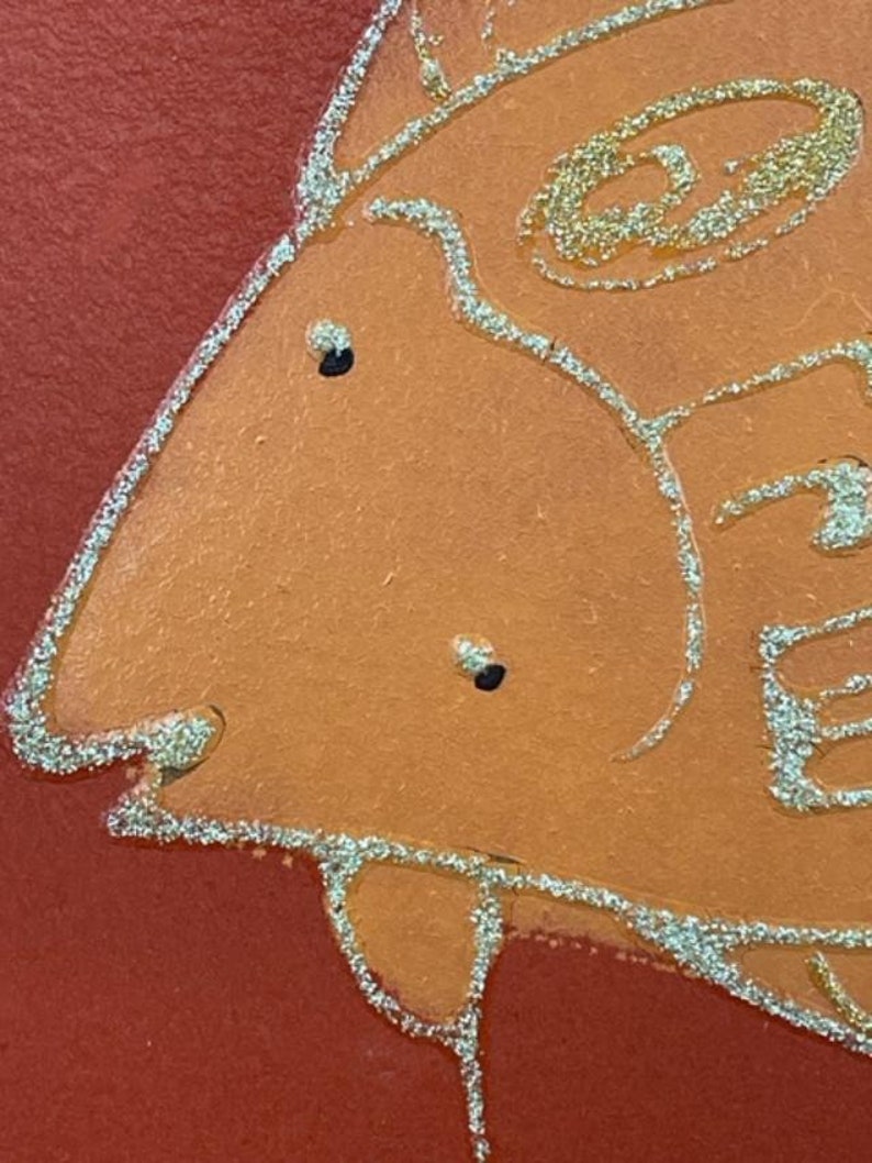 May include: Close-up of a stylized orange fish with gold glitter outlining the shape. The fish has two black eyes and a slightly open mouth.