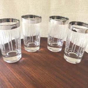 Puede incluir: Cuatro vasos altos transparentes con bordes plateados y líneas verticales grabadas, dispuestos sobre una bandeja de madera oscura. Los vasos están vacíos, ofreciendo un aspecto clásico y elegante.