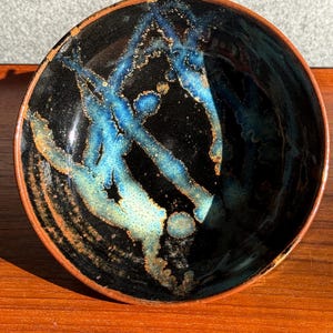 May include: A handmade ceramic bowl with a glossy black interior and a brown rim. The interior features striking blue and gold abstract patterns. The bowl is round and sits on a wooden surface.