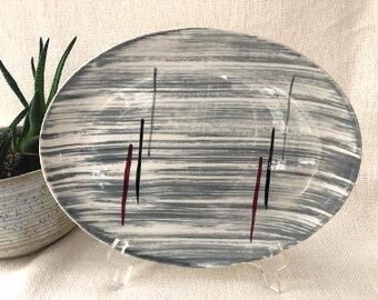 May include: A white oval serving platter with a gray and black abstract design. The design features thin, vertical lines and a few red accents.