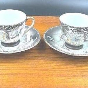 Puede incluir: Dos tazas y platillos a juego. Las tazas y los platillos son negros con un diseño de dragón blanco y dorado. Las tazas tienen asas doradas. Los platillos tienen un borde festoneado.