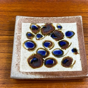 May include: Square ceramic dish with a brown rim and a white center. The center is decorated with a pattern of blue and brown circles.