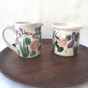 May include: Two ceramic containers with floral designs. The larger container is a cream-coloured pitcher with a handle. The smaller container is a cream-coloured jar. Both containers have a floral design featuring pink, purple, and green flowers.