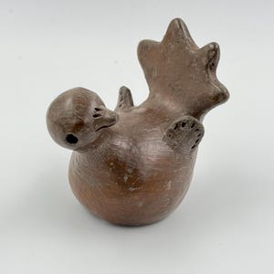 Puede incluir: Figura de ornitorrinco de cerámica marrón con la espalda arqueada y las patas en el aire. La escultura tiene un cuerpo redondeado, una cabeza pequeña con pico y una cola en forma de estrella. La figura está sobre un fondo blanco.